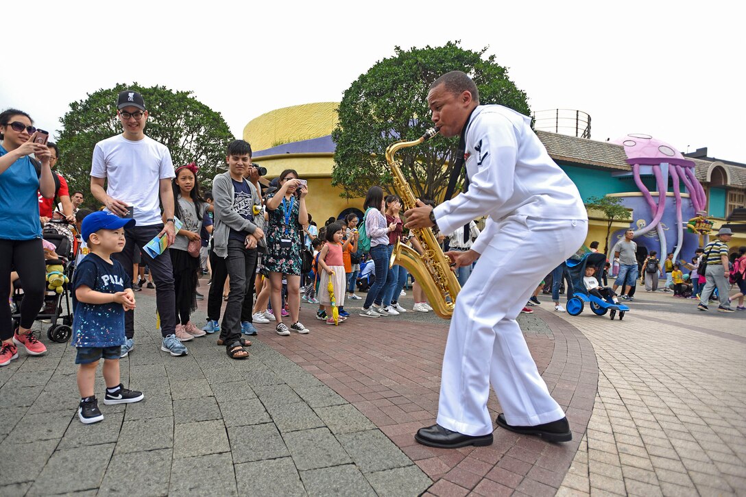 A sailor plays a saxophone at a theme park as a child watches.