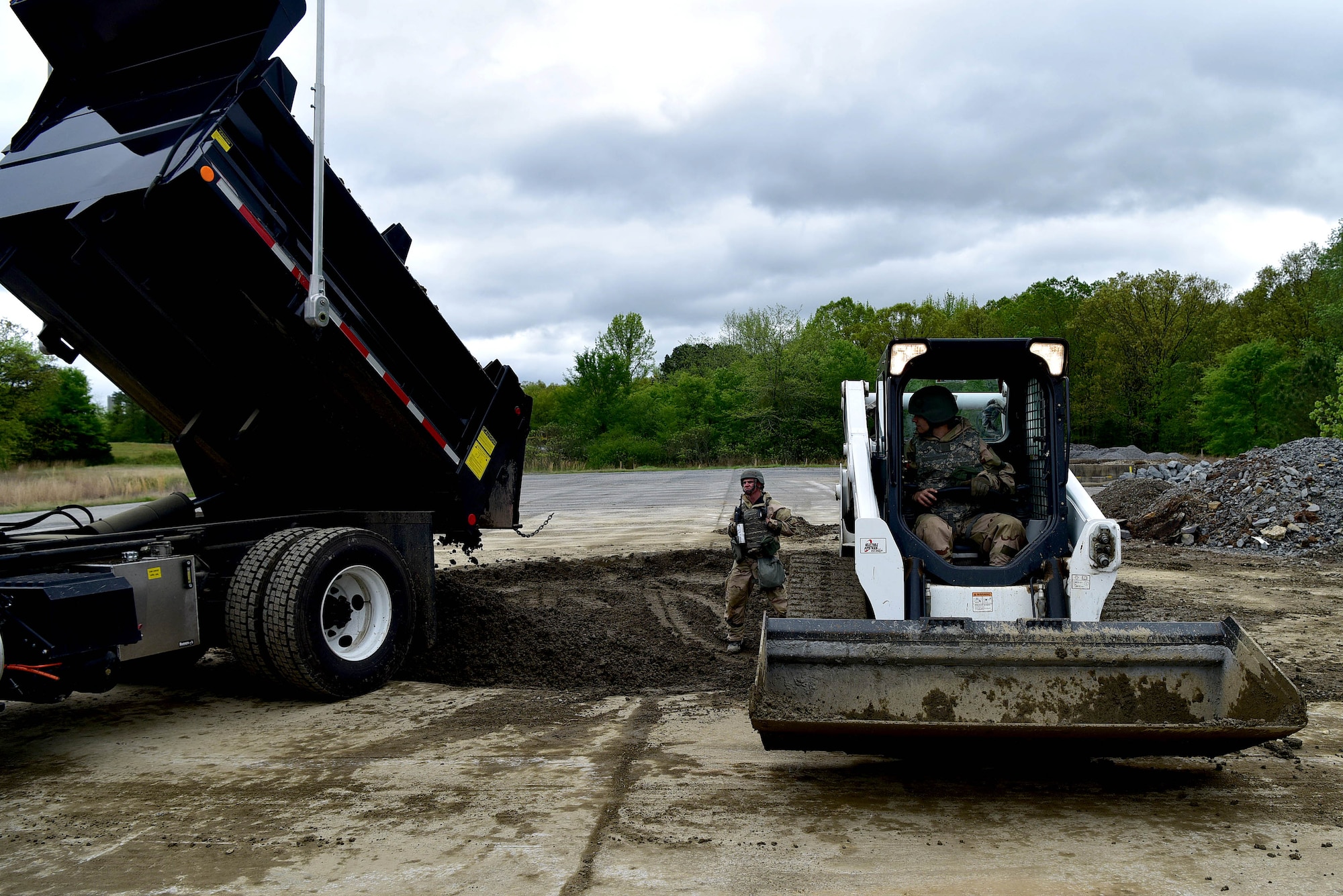 A man stands between two pieces of machinery as one dumps dirt and other minerals into a hole in the ground.