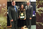 Three people pose for picture, middle person holds award.