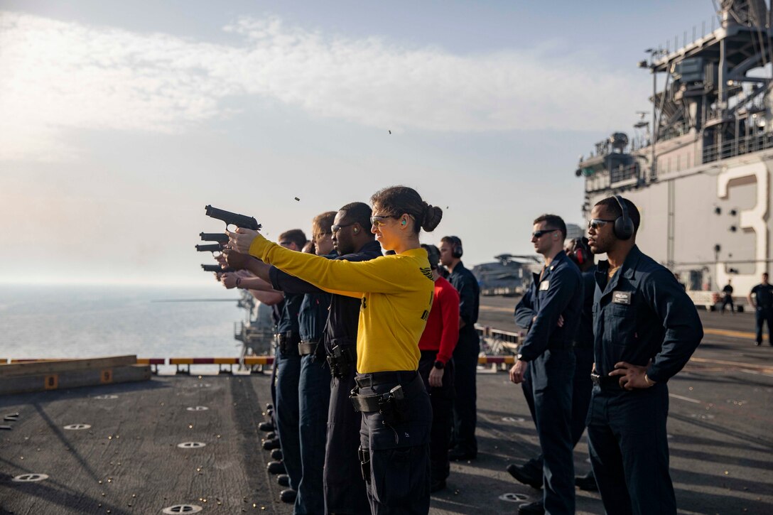 A female sailor in yellow and a line of sailors beside her fire pistols from a ship as instructors look on.