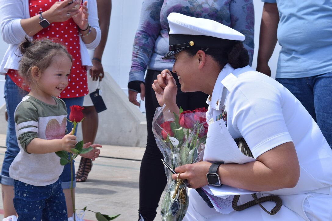 A girl holds a rose and smiles as she greets a sailor returning home.