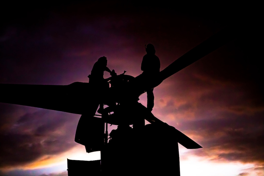 Two Marines are silhouetted against the twilight sky as they stand near the rotor of an Osprey performing maintenance.