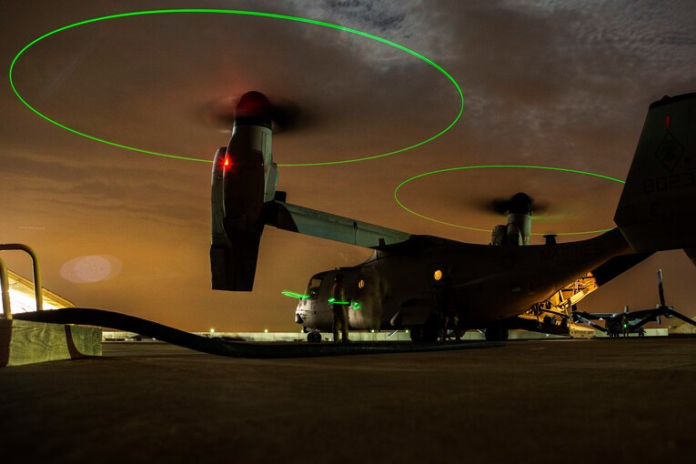 Two Marines check an Osprey at night as the rotors above them are outlined in green light.