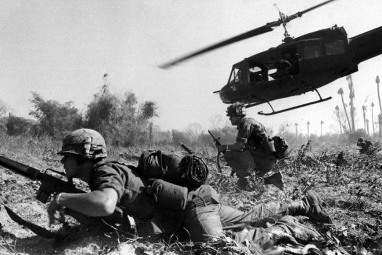 Two soldiers get in defensive positions on a field in Vietnam as a helicopter lifts into the air in the background.