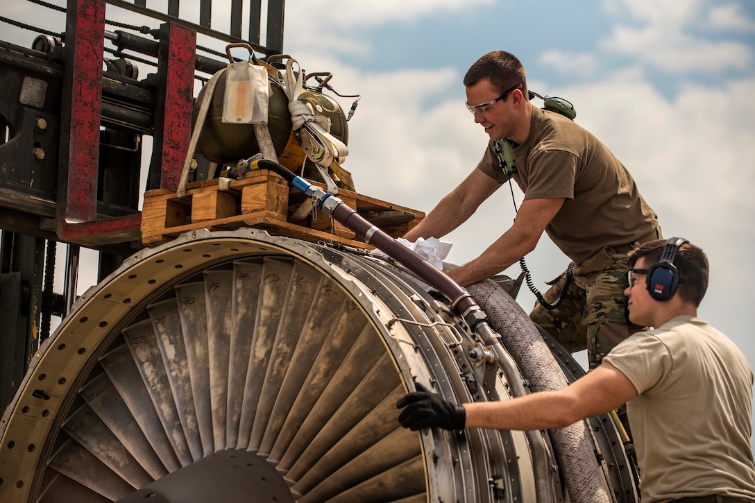 An airman secures an aircraft engine to a forklift as a second airman steadies the engine.