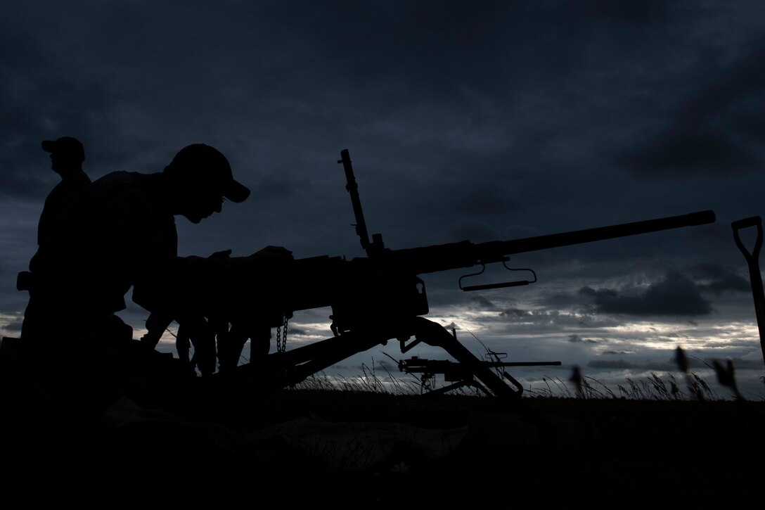 A man in a ballcap assembles a large machine gun in silhouette against the night sky as another man faces the opposite direction.