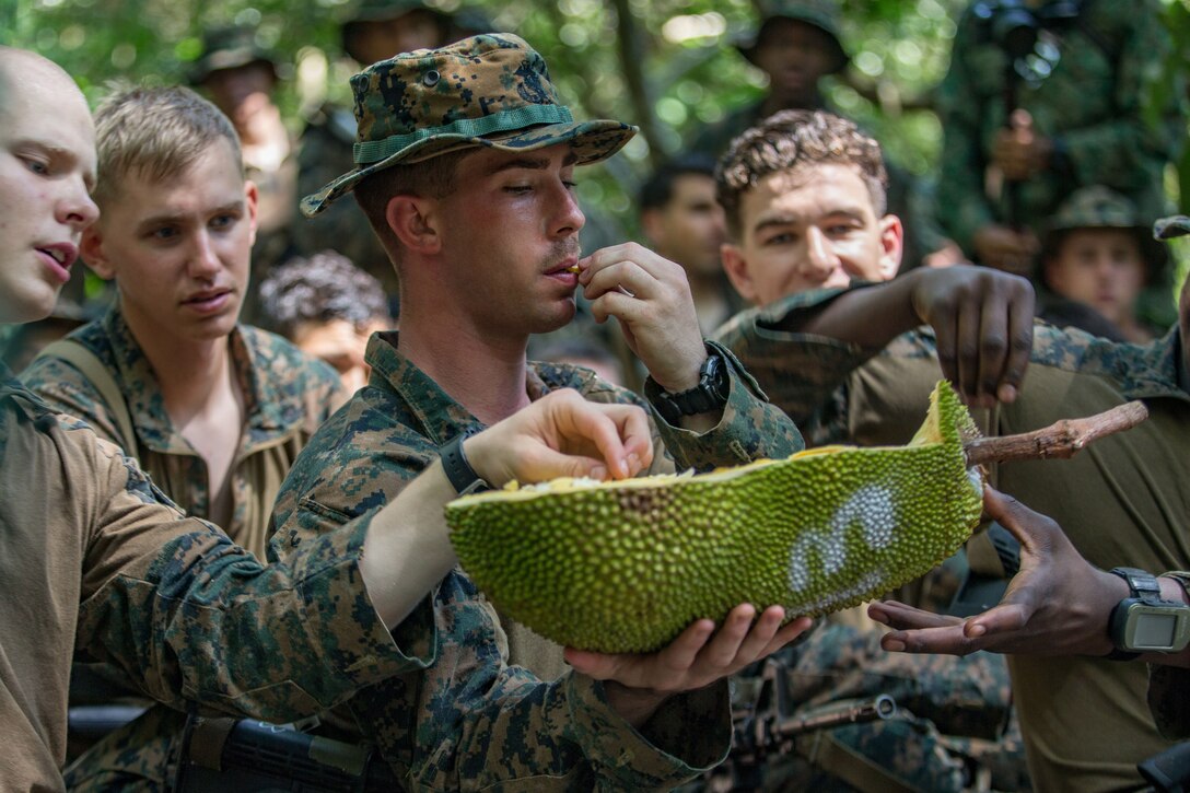 A Marine wearing a cover holds a jackfruit and tastes it as other Marines reach in for a piece of it.