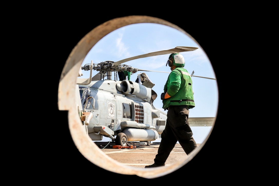 Two sailors work on top of a helicopter as another sailor walks by.