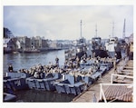 Color photo of troops in Landing Crafts with LCI(L)-84 and other naval vessels in the background.