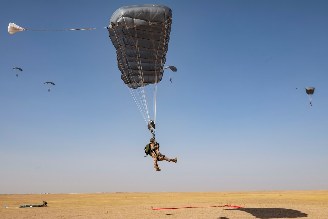 Two Marines, shown from the side with a parachutes above them, descend through the sky.