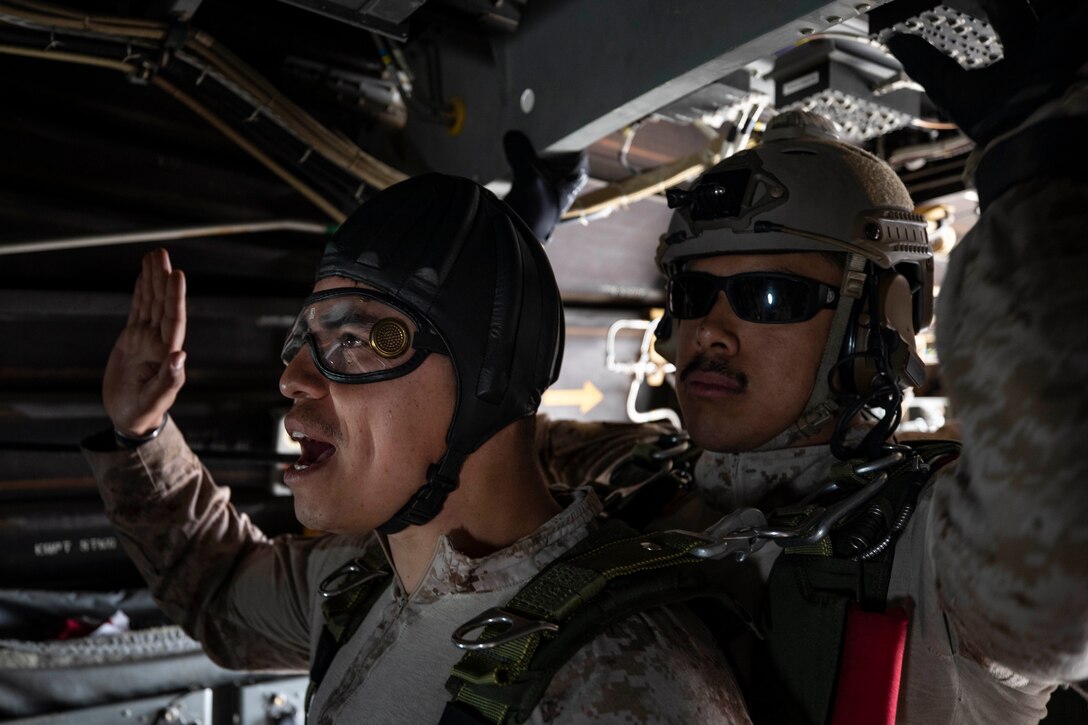 A man wearing headgear and goggles raises his hand while a second man attached to him steadies himself near the open door of an aircraft.