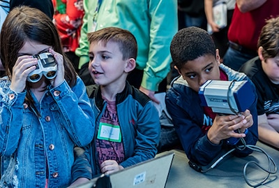 Girl looking through binoculars, boy watching girl and another boy holding a radar gun.