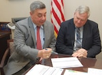 Two men wearing business suits sit at a table discussing a briefing