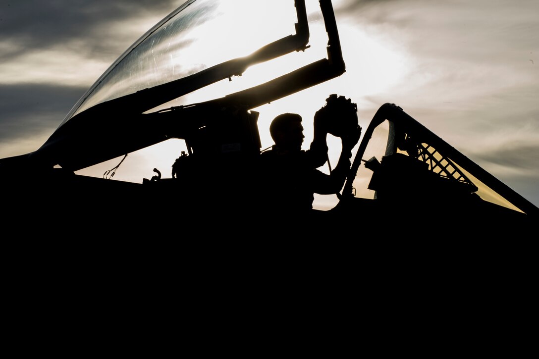 A pilot puts on his helmet while sitting in a fighter jet at twilight.