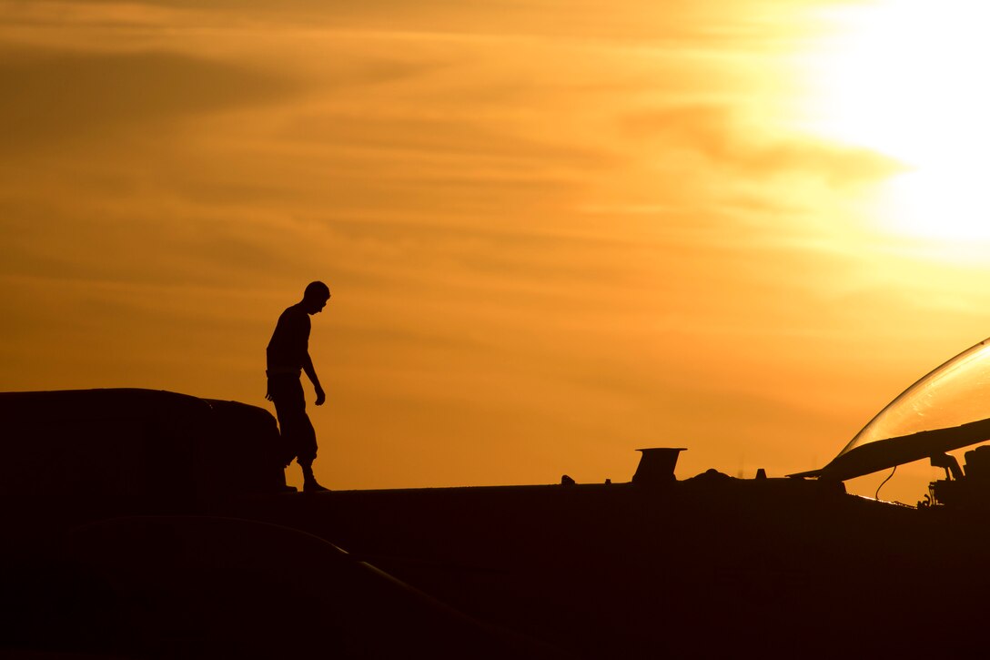 An airman walks on top of a fighter jet  at sunset.