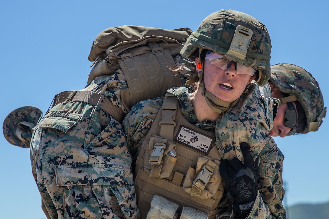 A Marine wearing protective lenses and a helmet carries another Marine across her shoulders.