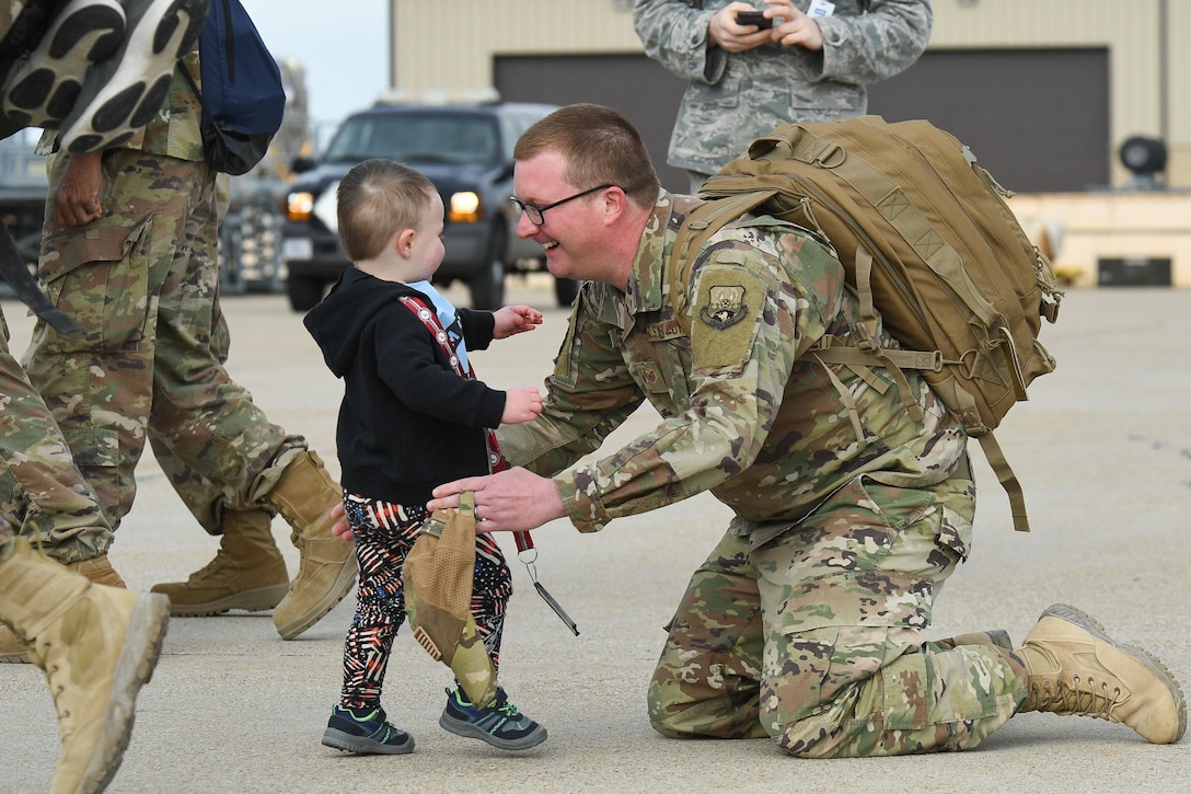 A man wearing a uniform and backpack kneels on pavement and reaches out to greet a toddler who has walked up to him.