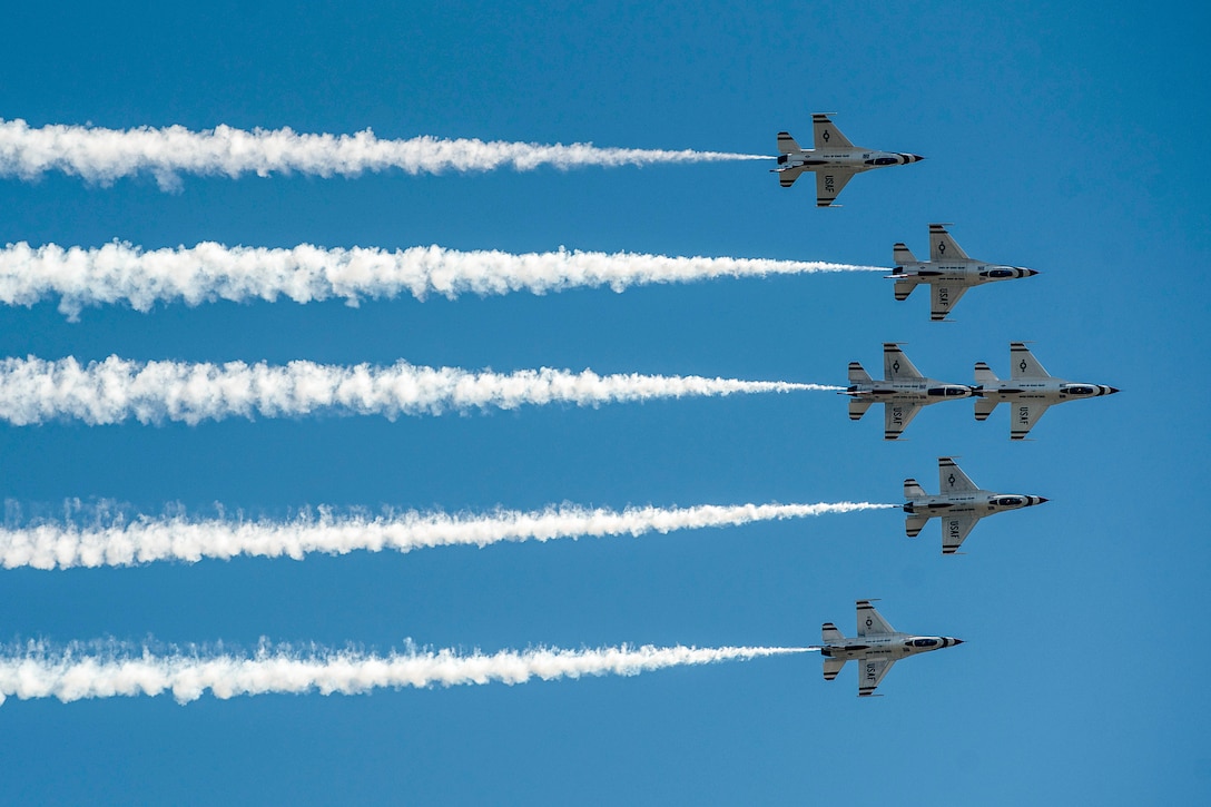 Six jets fly in blue sky in a triangular formation, leaving white smoke trails in their wake.