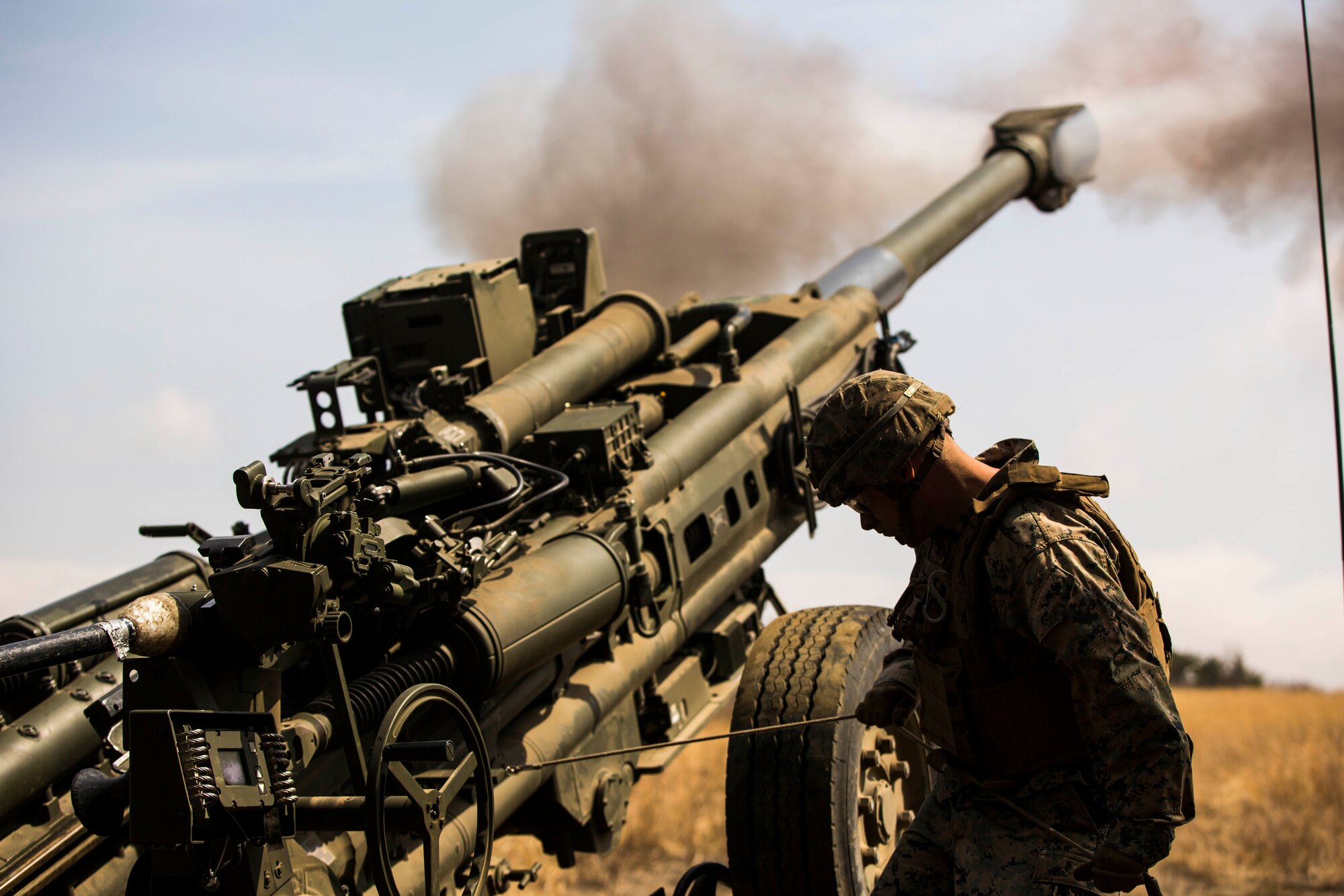A Marine stands next to a large military gun that has smoke coming out from the top.