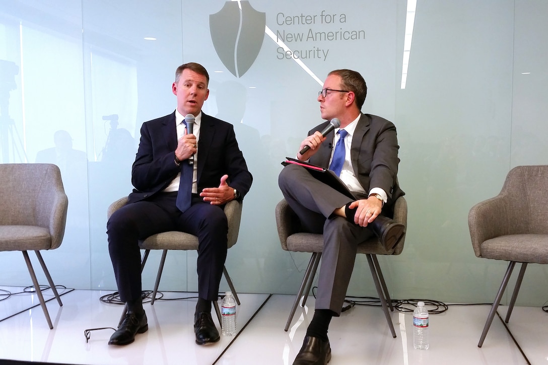 Two men speak to each other, seated on a podium.