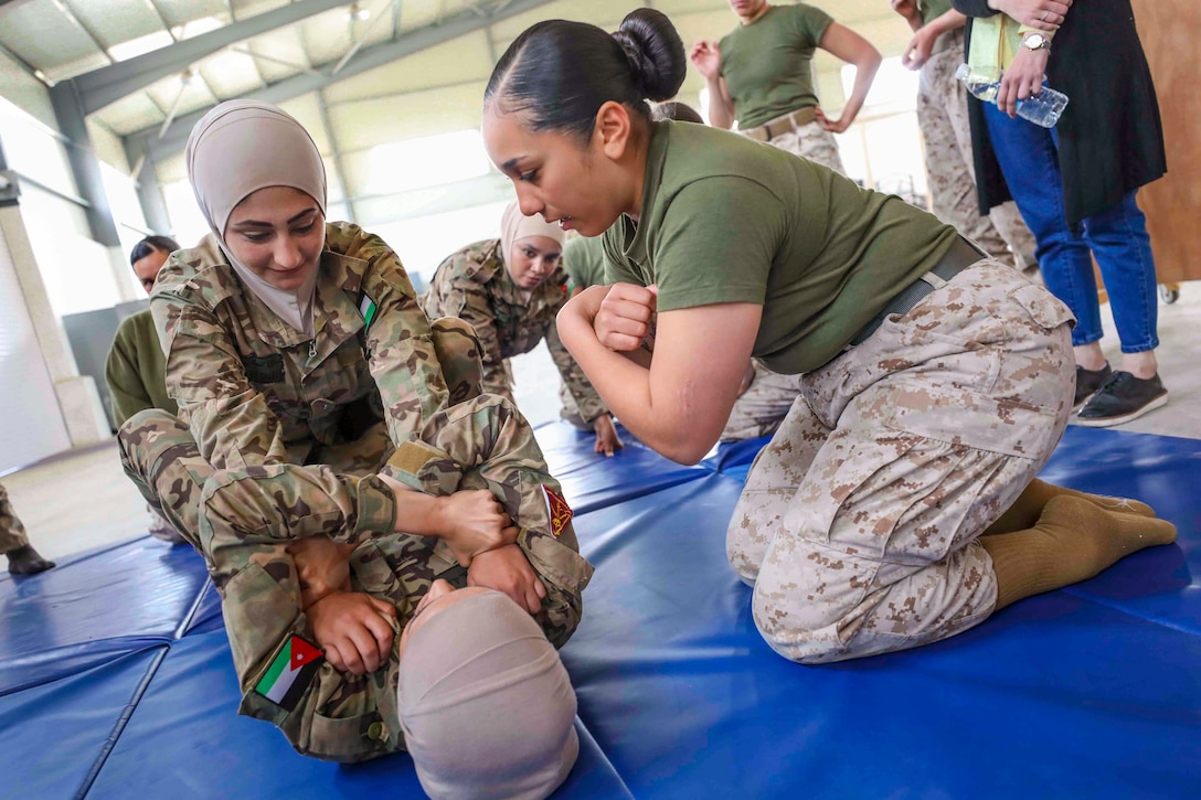 A Marine kneels on the ground next to two other soldiers who are wrestling.