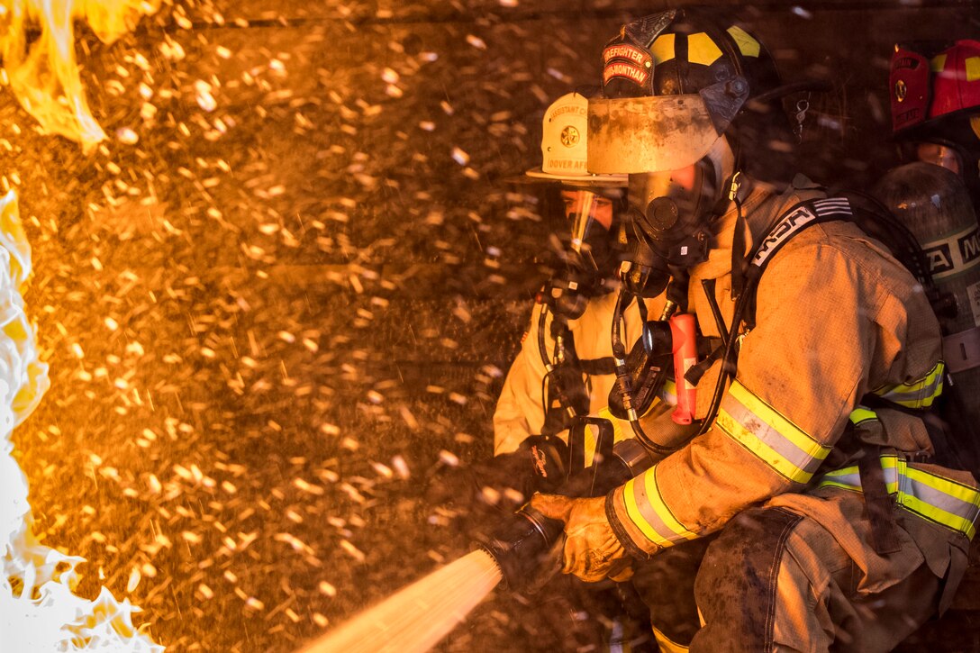 A firefighter aims water from a fire hose at flames as another firefighter monitors.