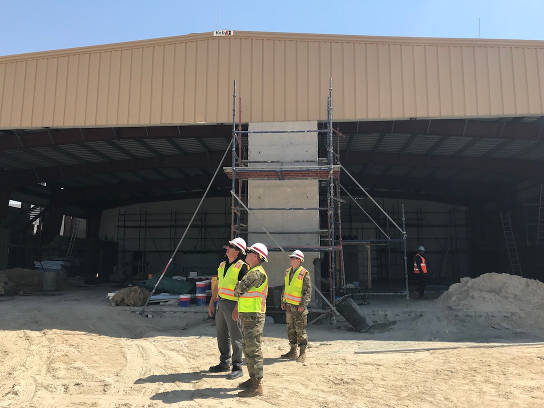 3 people in military uniform standing in front of a large building under construction. They are standing on sand and the front of the building is open.