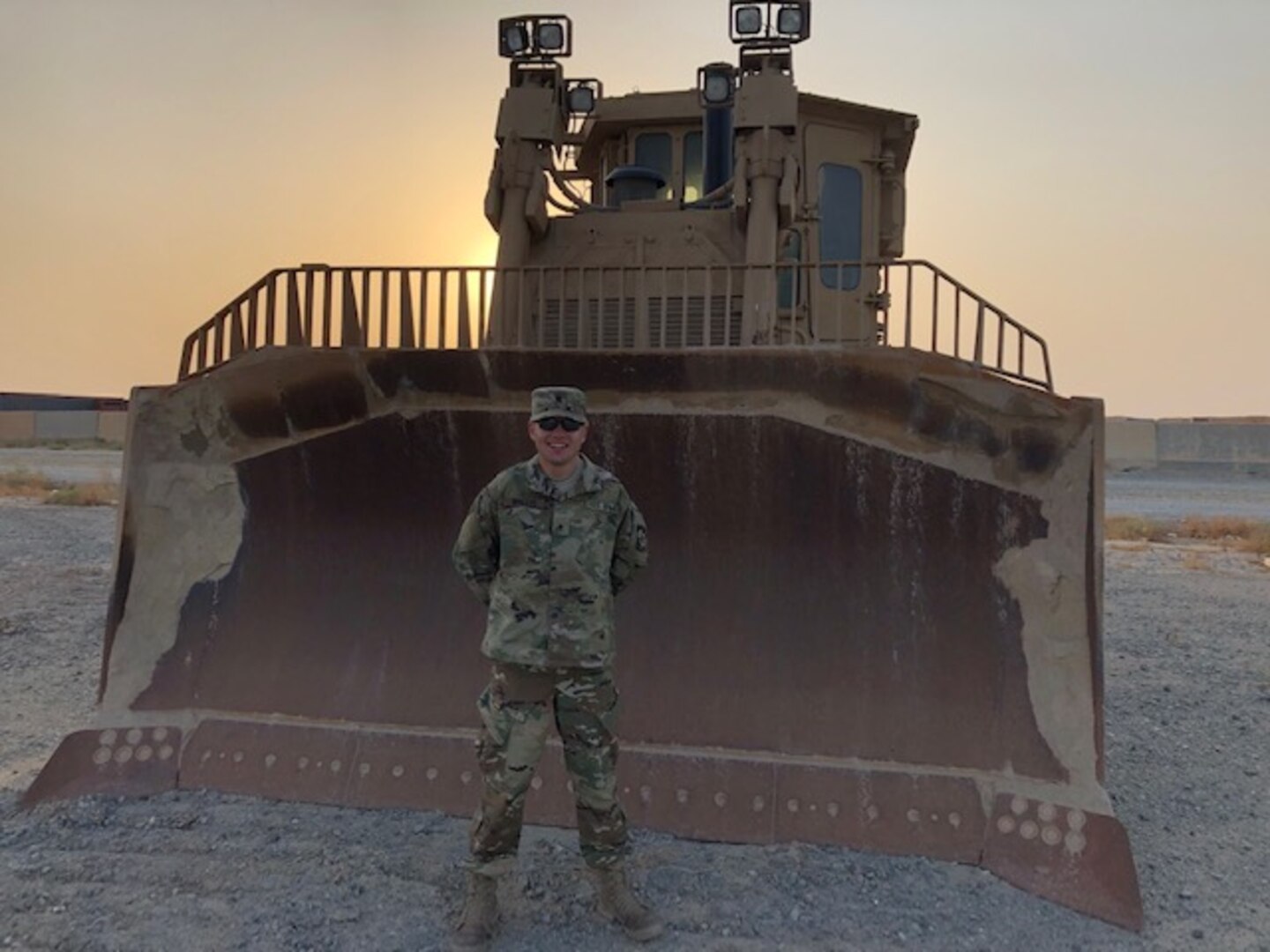 Man in military uniform standing in front of a large bull dozer in the desert.