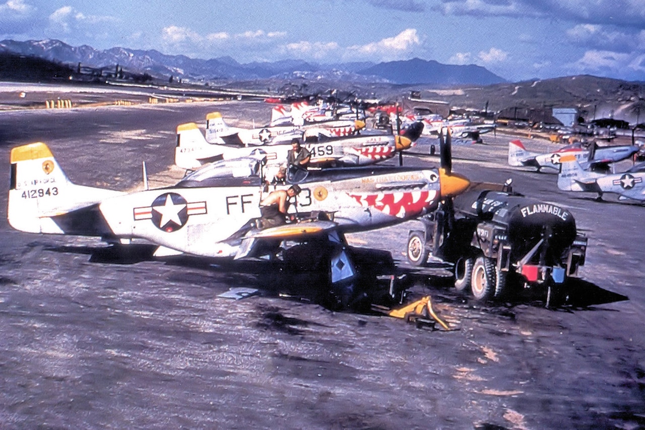 Aviation mechanics work on a runway full of painted F-51 Mustang airplanes. Beside the closest airplane is a black tanker with the word “flammable” on it.