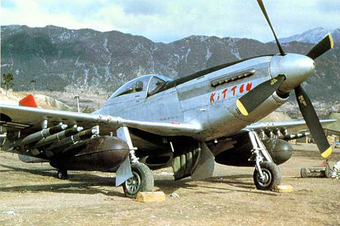 An airplane with the word “kitten” written on the side sits on gravel with mountains in the background. The plane has a propeller and has bombs and rockets strapped underneath its wings.