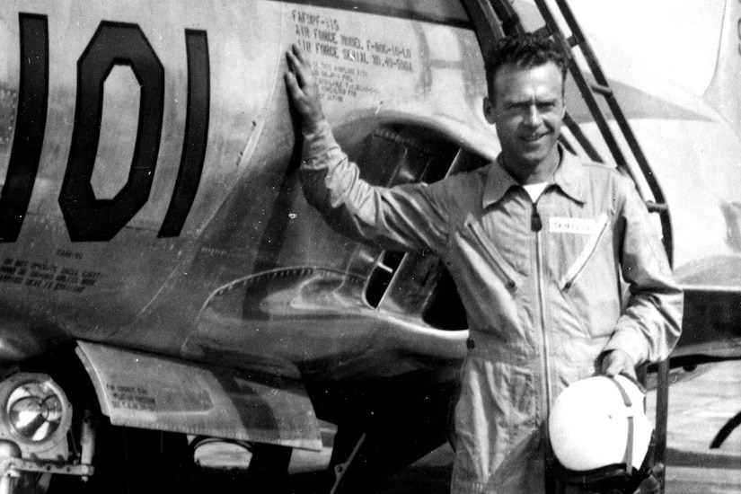 A pilot in a flight suit holds his helmet in one hand and props himself up against a fighter jet with the other. Stairs up to the plane’s cockpit are behind him.