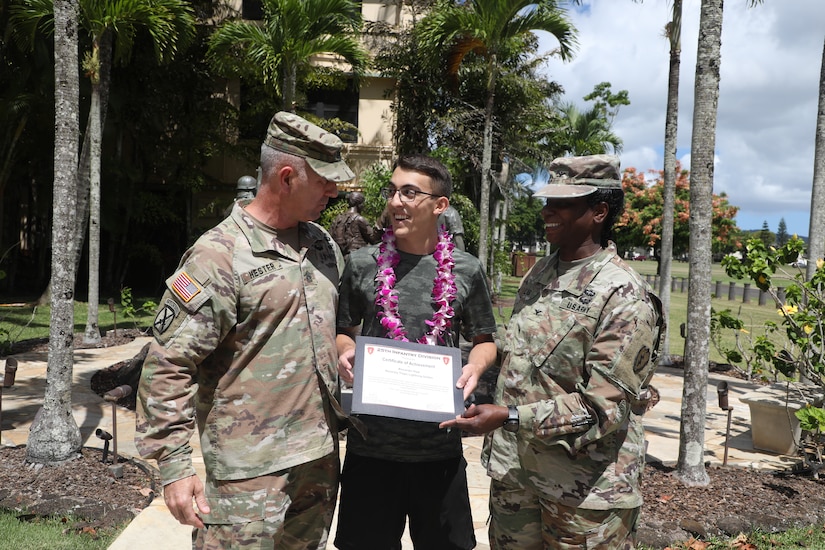 A teen accepts a certificate from two Army leaders. A statue and several palm trees are in the background.