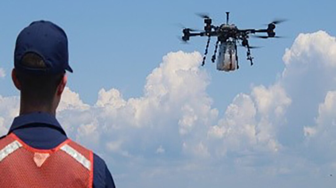 A man in a reflective vest looks on as a drone carrying a test payload flies over the ocean