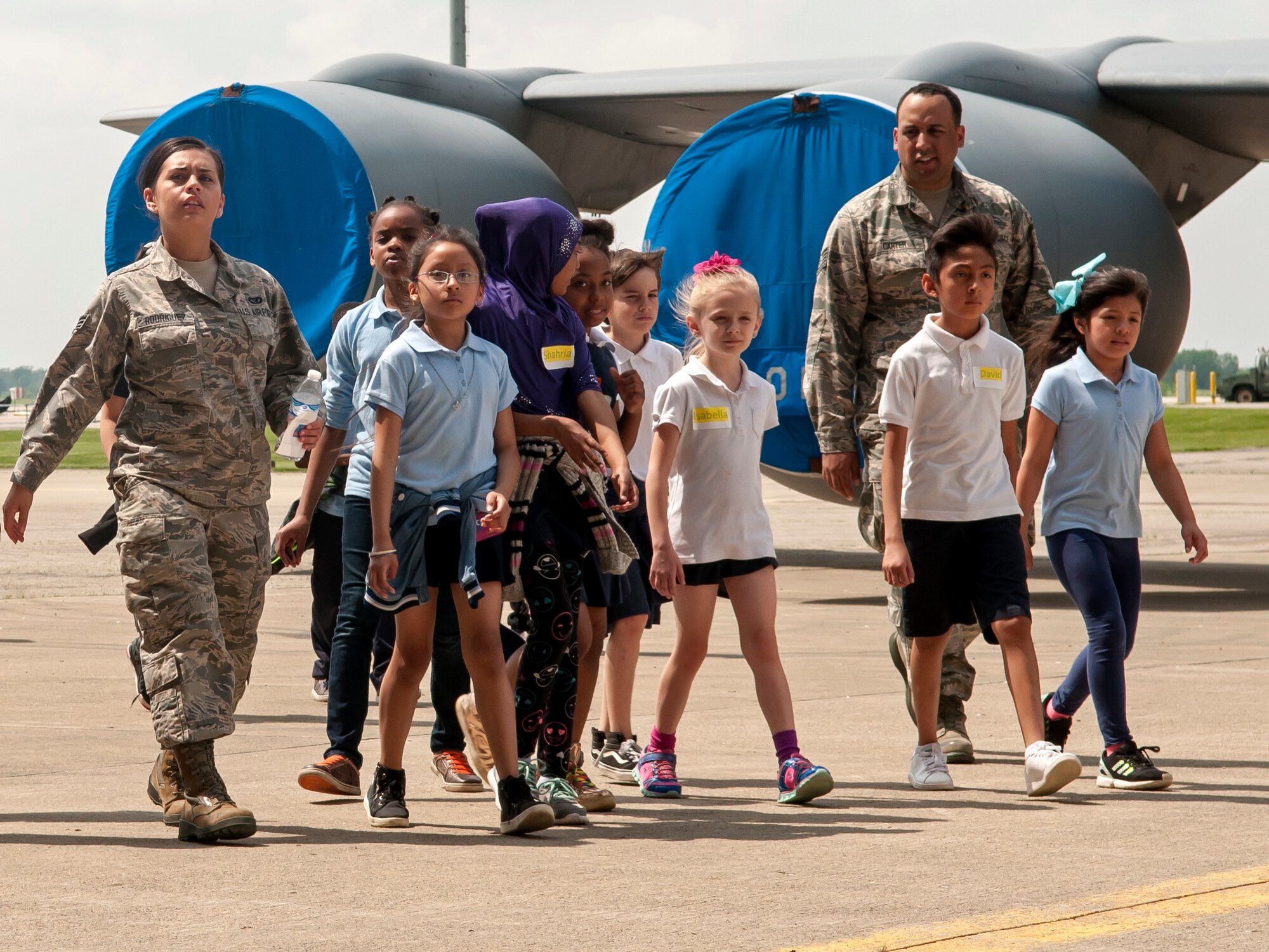 Two Airmen with  a group of children walking on the flight line with a plane visible in the background