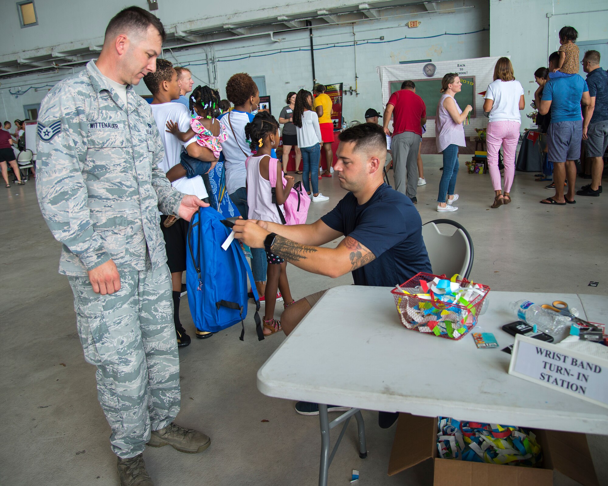 U.S. Air Force Senior Airman Jason Wisman, a 6th Aircraft Maintenance Squadron crew chief, cuts a wrist band off a backpack for U.S Air Force Staff Sgt. David Wittenauer, a 927th Security Forces Squadron defender, at a Back to School Info Fair hosted by the 6th Force Support Squadron at MacDill Air Force Base, Fla., Aug. 3, 2019.