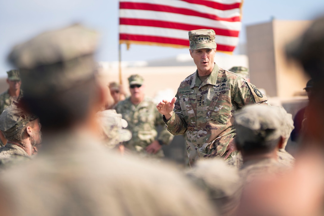 An Army officer in camouflage uniform speaks to seated men with an American flag in the background.