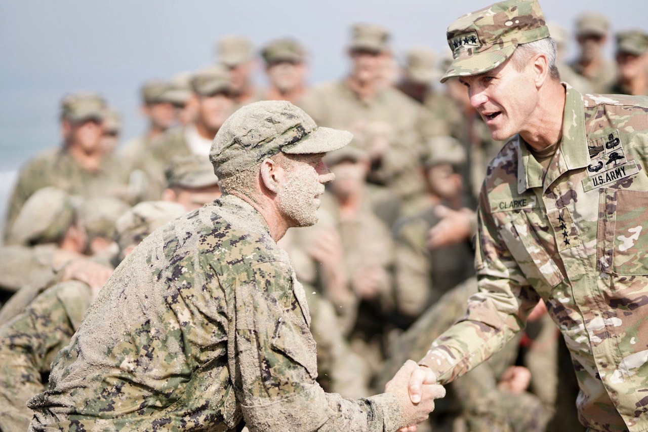 An Army officer shakes hands with a sand-covered sailor. Both are in camouflage uniforms.