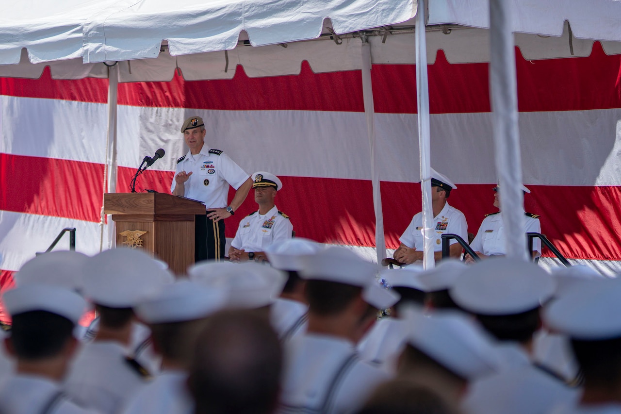 An Army officer in dress uniform and tan beret speaks to seated sailors wearing “Cracker Jack” uniforms.