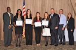 A group of people stand in front of a room. Four women are holding awards.