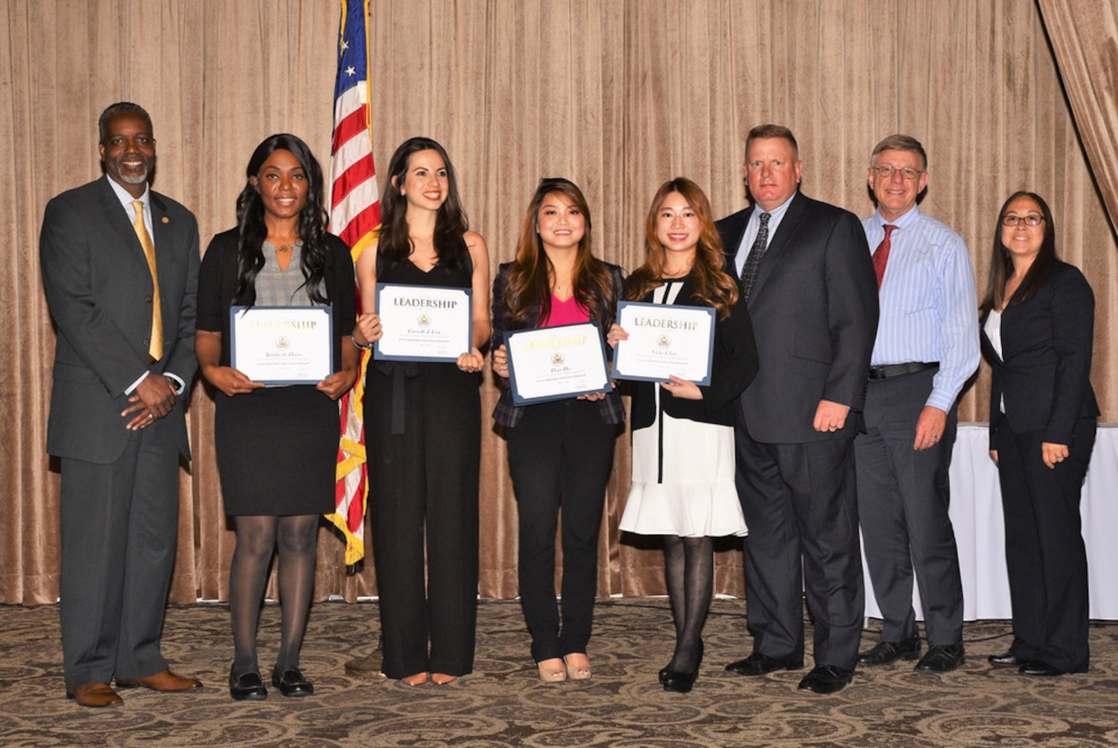 A group of people stand in front of a room. Four women are holding awards.