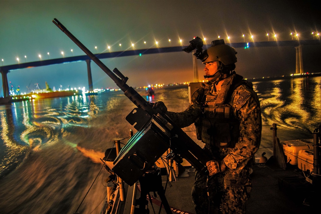 A sailor mans a machine gun aboard a boat in water at night, with a bridge in the background.