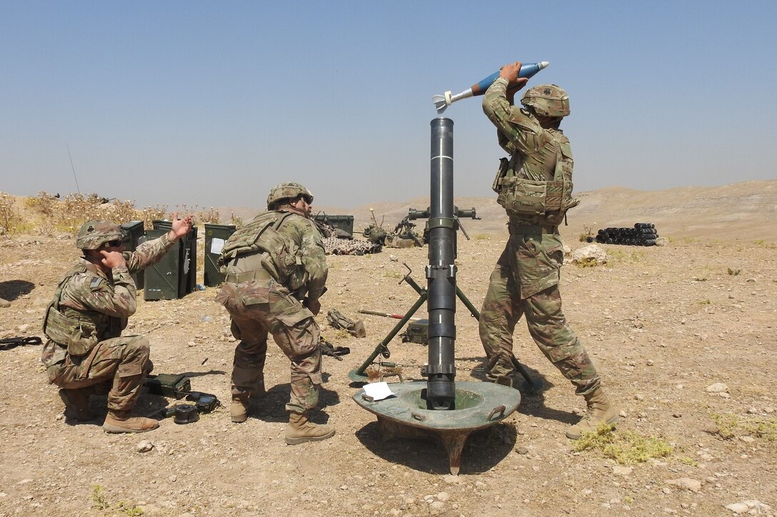 A soldier loads a round into a mortar as others crouch nearby in desert-type terrain.