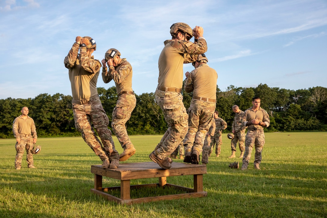 Four service members jump off a low wood platform in a field.