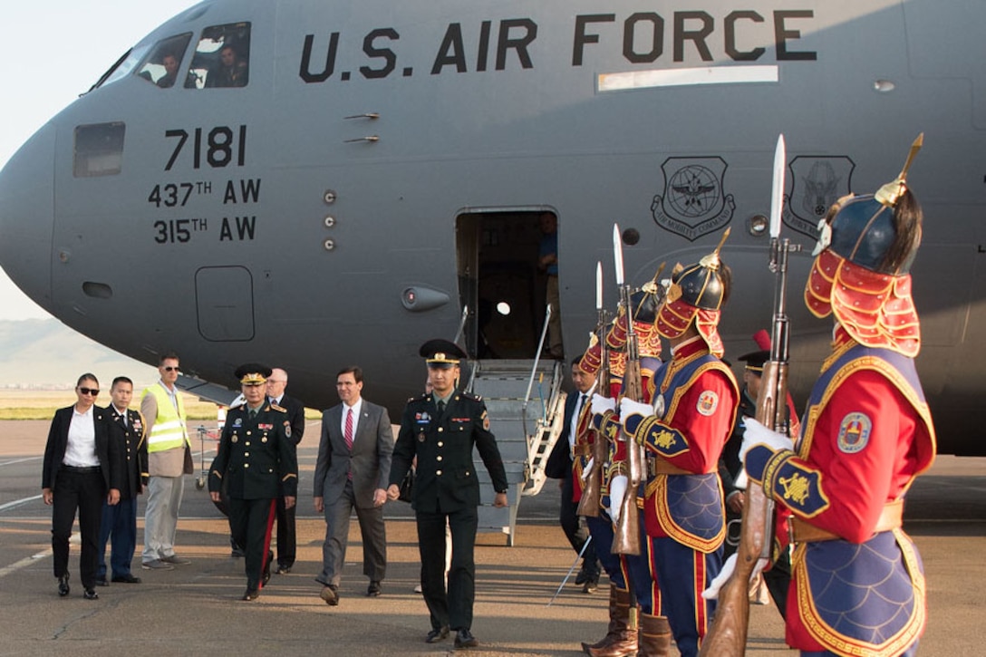 Defense Secretary Dr. Mark T. Esper walks alongside a group of people with a plane in the background.