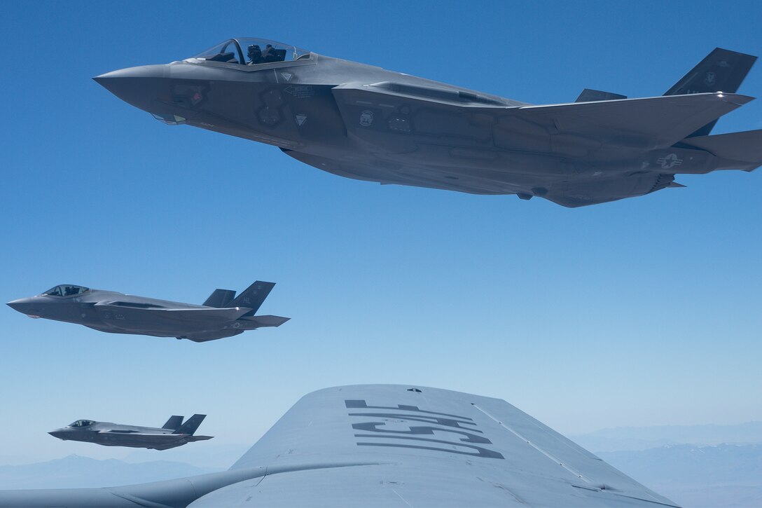 Three military fighter jets are seen off the wing of another military aircraft.