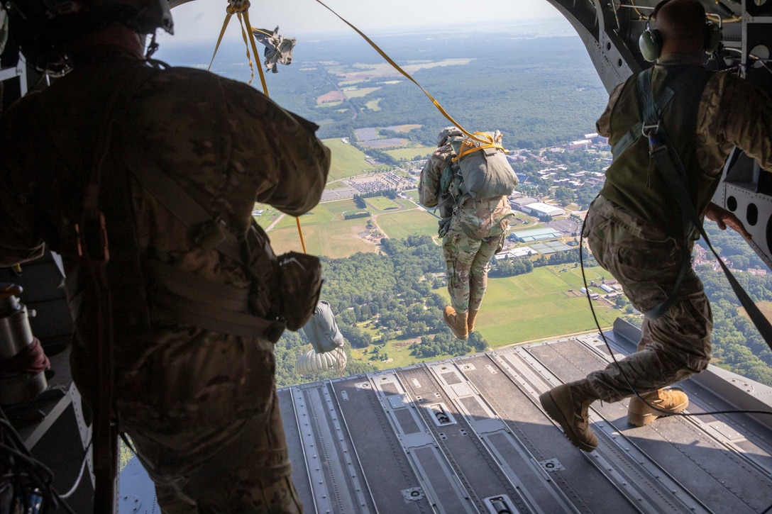A soldier jumps out of plane while two other soldiers stand on either side inside the plane.