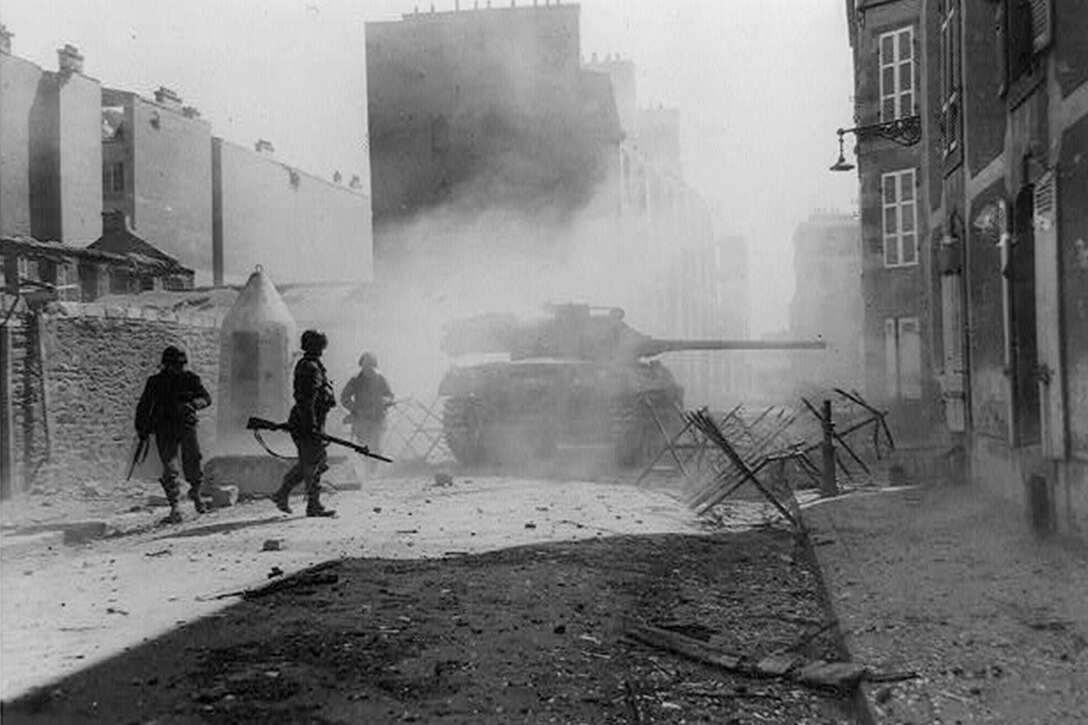 Three soldiers with rifles walk beside a tank along a tattered street in a town surrounded by several buildings.