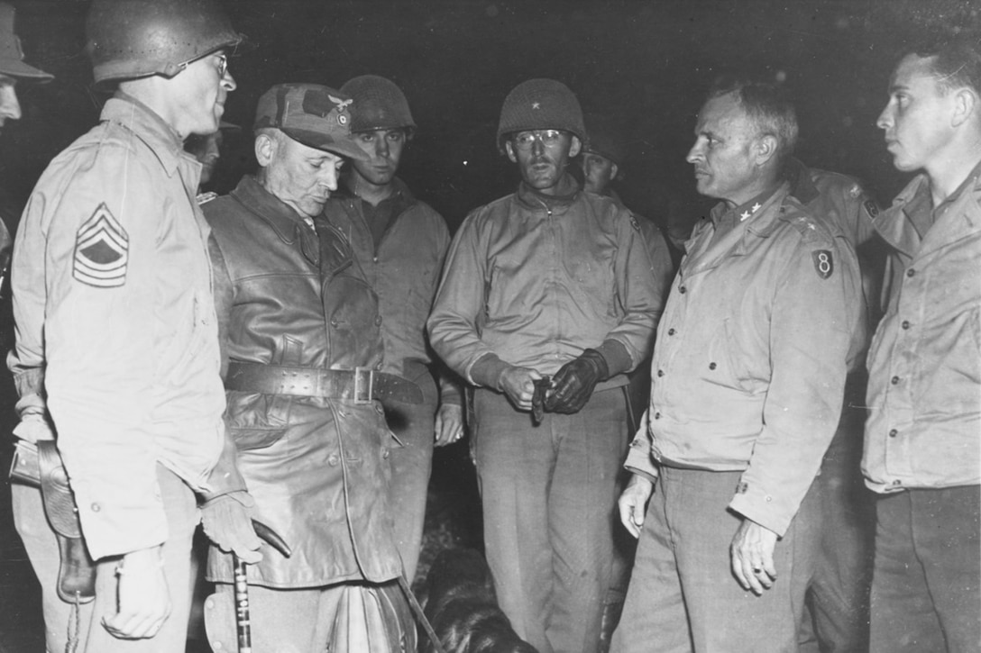 Several American soldiers wearing Army Eighth Division patches stand with a German general and his dog.
