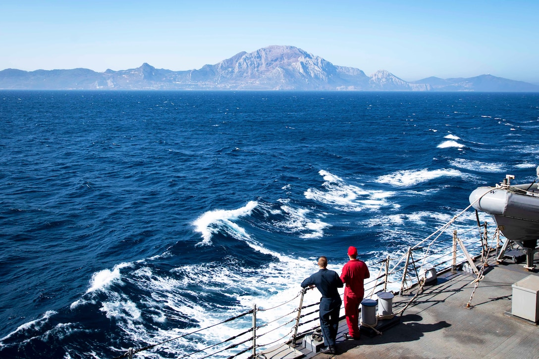 Two sailors on a ship look at waves and Gibraltar.