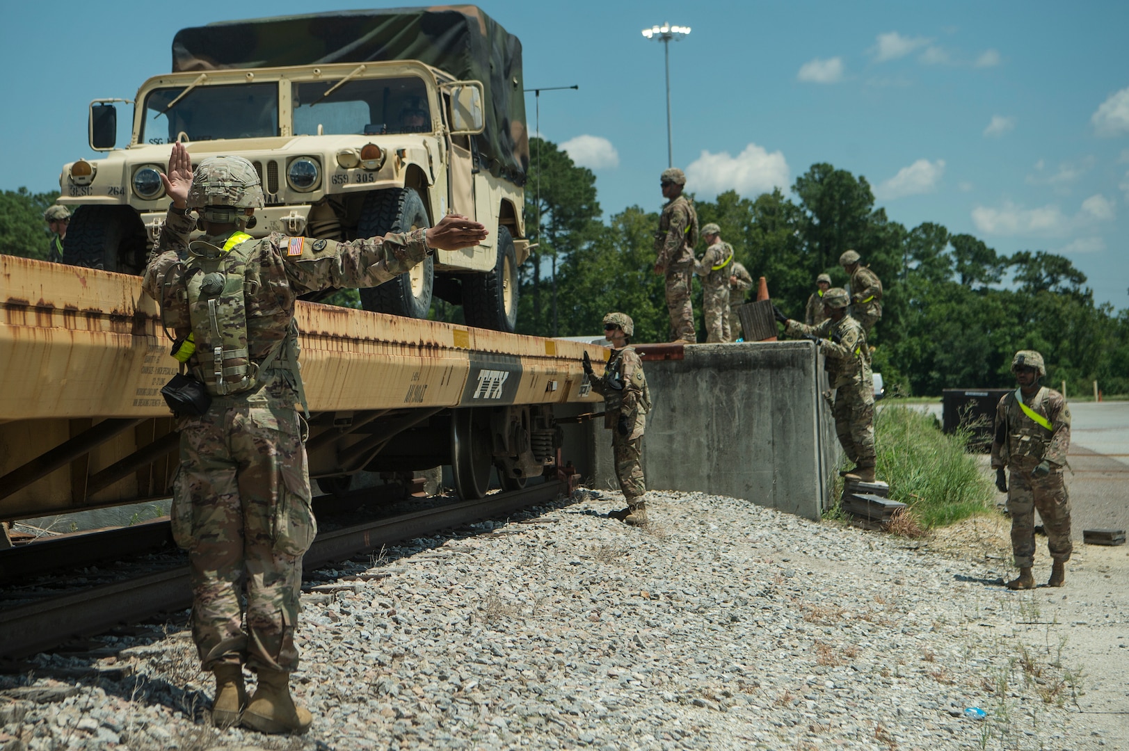 A soldier in camouflage uniform and wearing a helmet uses arm signals to guide a tactical truck onto a yellow flatbed railcar.